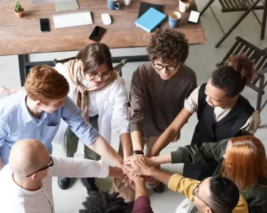 A diverse group of professionals joins hands in a team-building exercise indoors.
