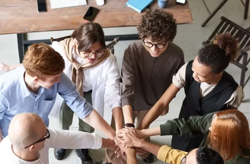 A diverse group of professionals joins hands in a team-building exercise indoors.
