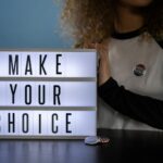 Woman holding lightbox with 'Make Your Choice' promoting civic duty and voting.