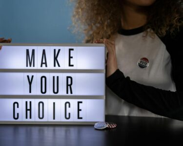 Woman holding lightbox with 'Make Your Choice' promoting civic duty and voting.