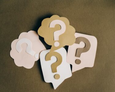 Four paper cutouts of question marks in beige and white on a brown surface, symbolizing inquiry and curiosity.
