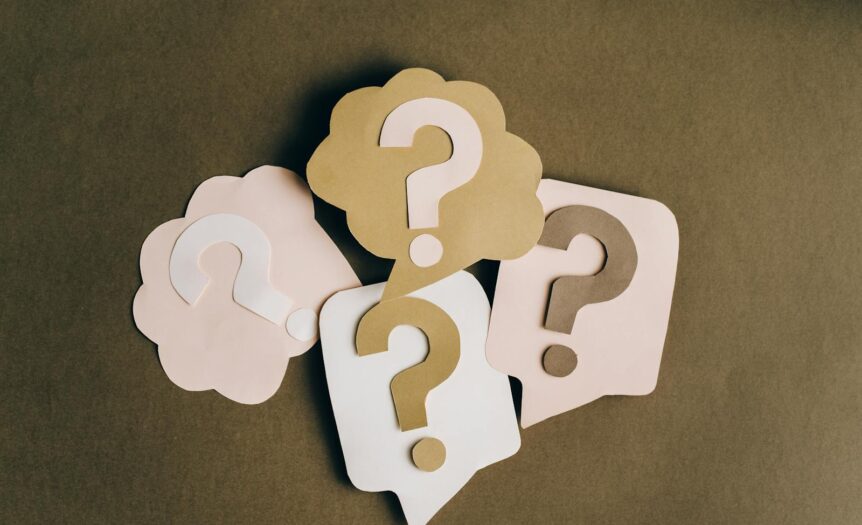 Four paper cutouts of question marks in beige and white on a brown surface, symbolizing inquiry and curiosity.