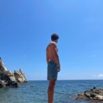 topless man in blue shorts standing on rock formation near body of water during daytime