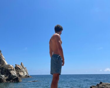 topless man in blue shorts standing on rock formation near body of water during daytime