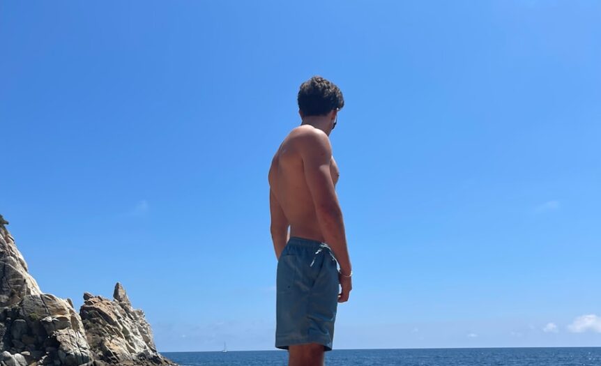topless man in blue shorts standing on rock formation near body of water during daytime