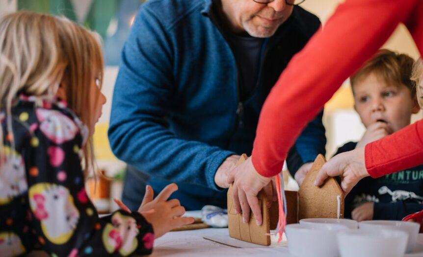 two person making gingerbread house and three children watching them