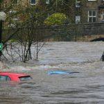 Three cars partially submerged in a flooded river near a stone bridge