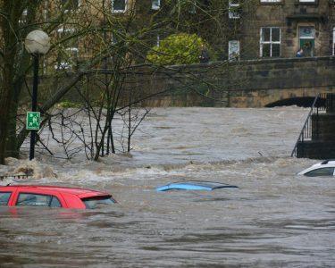 Three cars partially submerged in a flooded river near a stone bridge